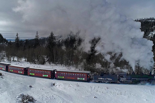 Steam train travels through snowy landscape with trees and mountains in background.