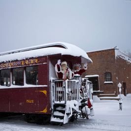 Santa waving from snowy red train car next to brick building in winter scene.