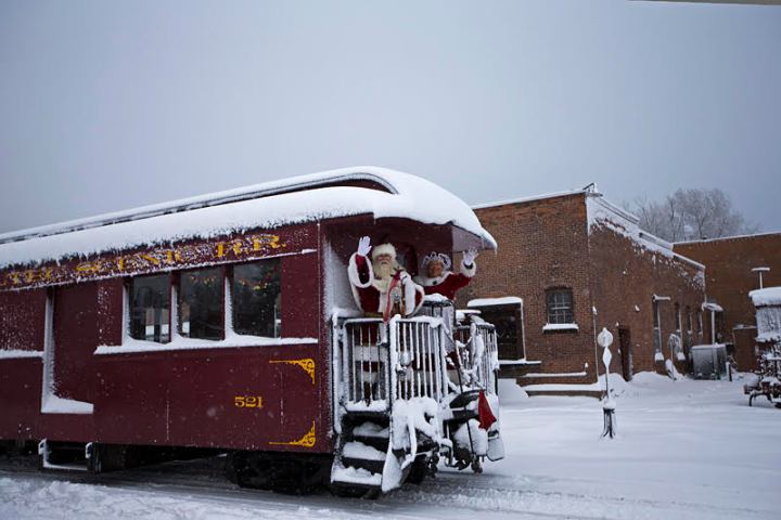 Santa waving from snowy red train car next to brick building in winter scene.