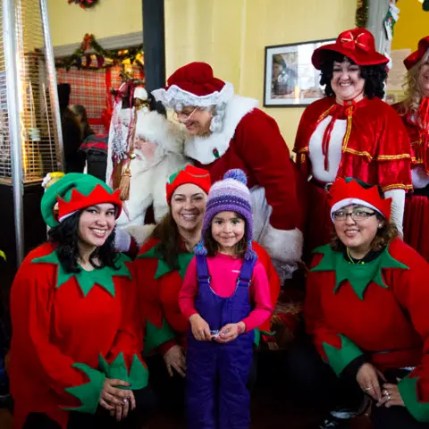 Group of people in festive costumes posing, including elves and Santa, with a young girl in center.