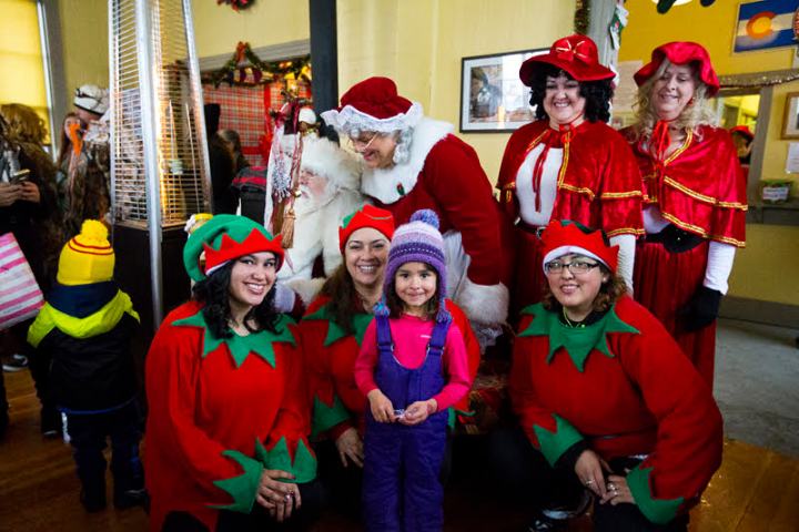 Group of people in festive costumes posing, including elves and Santa, with a young girl in center.