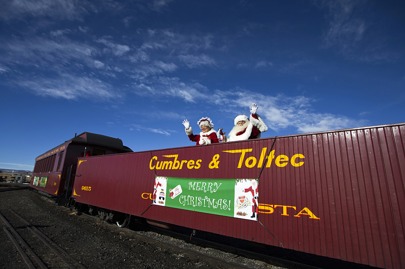 Santa and Mrs. Claus wave from Cumbres & Toltec train with 'Merry Christmas' sign.