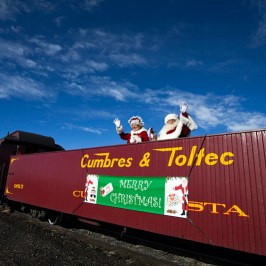 Santa and Mrs. Claus wave from Cumbres & Toltec train with 'Merry Christmas' sign.