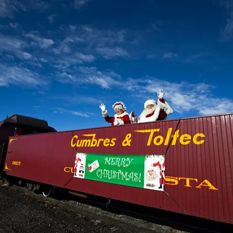 Santa and Mrs. Claus wave from Cumbres & Toltec train with 'Merry Christmas' sign.