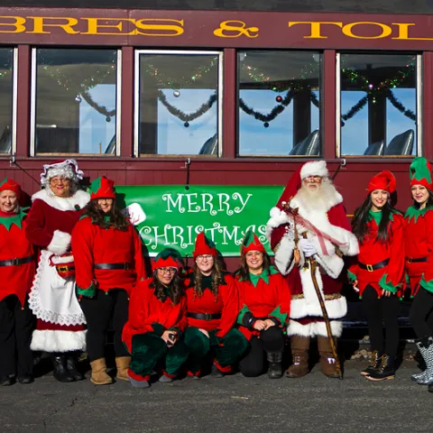 Santa and elves posing in front of a decorated train with a 'Merry Christmas' sign.