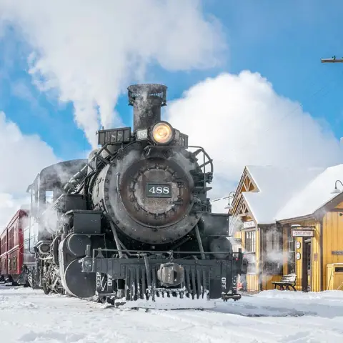 Steam train near a snowy station labeled 'Chama' with blue sky and clouds.