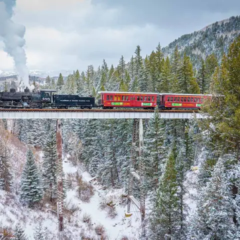 Steam train crosses snowy forest bridge, billowing smoke, vibrant passenger cars.