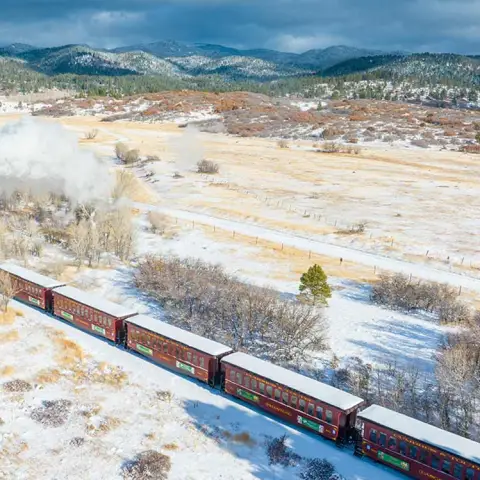 Steam train passing through snowy landscape with distant mountains.