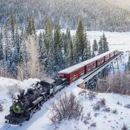 Steam train crosses snowy bridge in a forested winter landscape.
