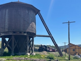 Old water tower, vintage train, and yellow building under clear blue sky.
