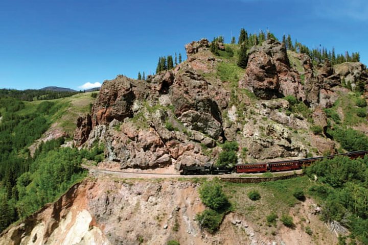 Train winding through rocky green hillside under clear blue sky.