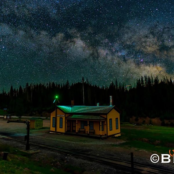 Starry night sky over a small yellow building with trees in the background.