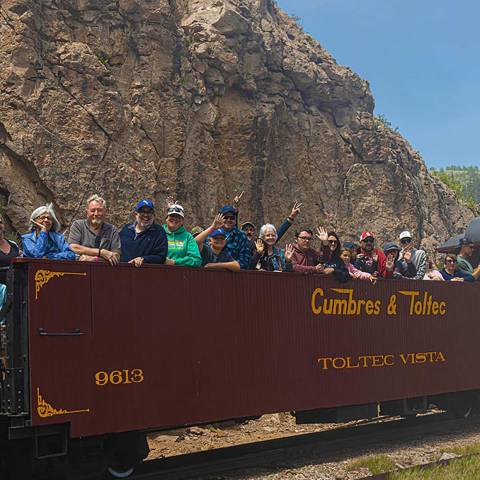 People riding on an open train carriage labeled Cumbres & Toltec, with rocky cliffs in the background.