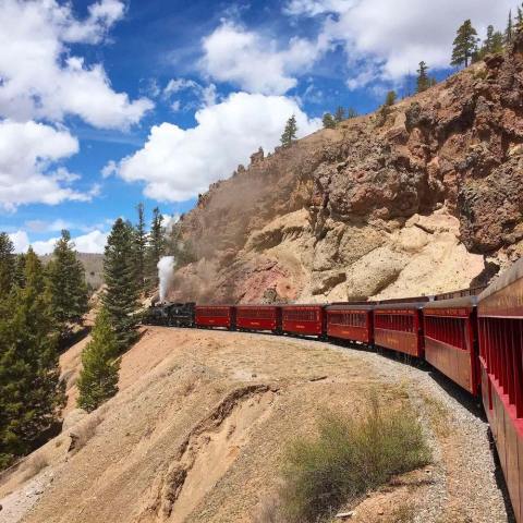 Red train on a mountainside with pine trees and blue sky with clouds.