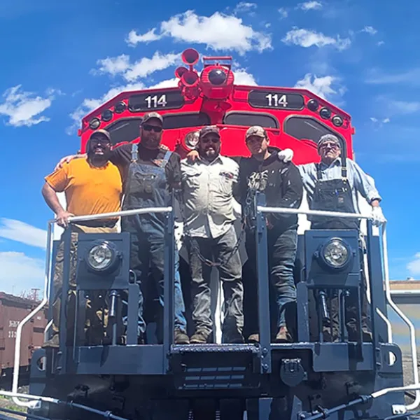 Train crew standing on locomotive 114 smiling under a clear blue sky.