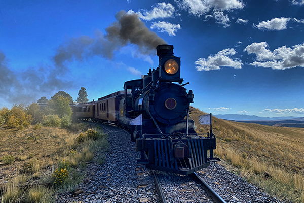 Steam train traveling on tracks through open countryside under a clear blue sky.