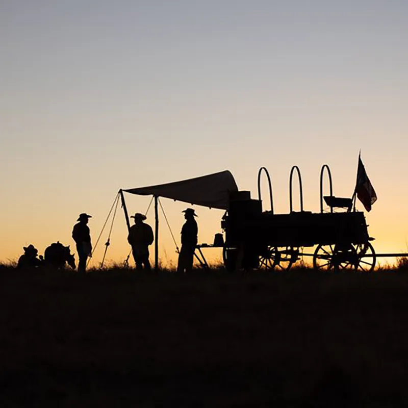 Silhouetted cowboys and covered wagon at sunset.