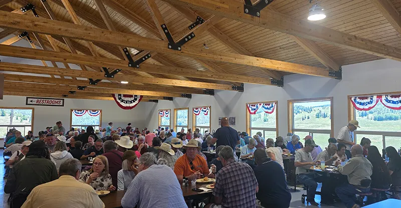 Crowded dining hall with people sitting at tables under wood beam ceiling.