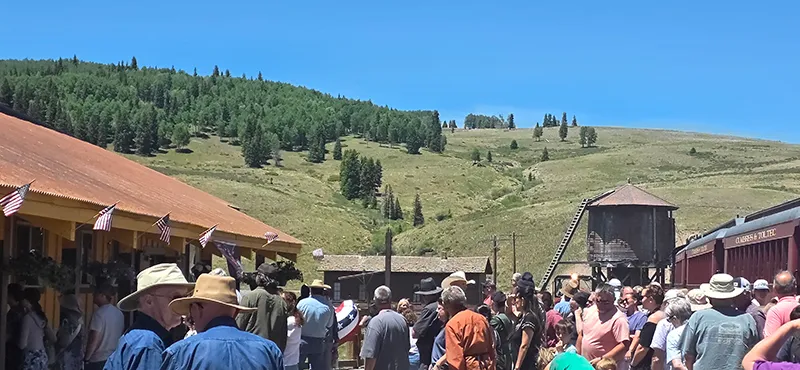 Large crowd gathered at historic Osier Station in a grassy rural area with a water tower and hills in the background.