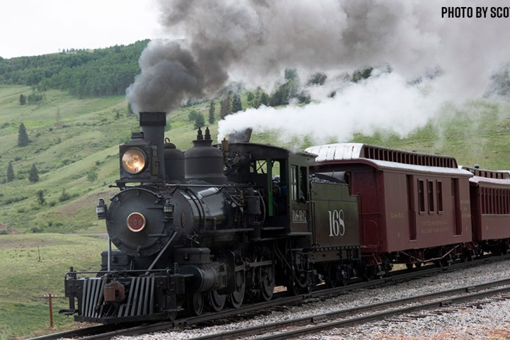 Steam locomotive on tracks in rural landscape with smoke billowing.