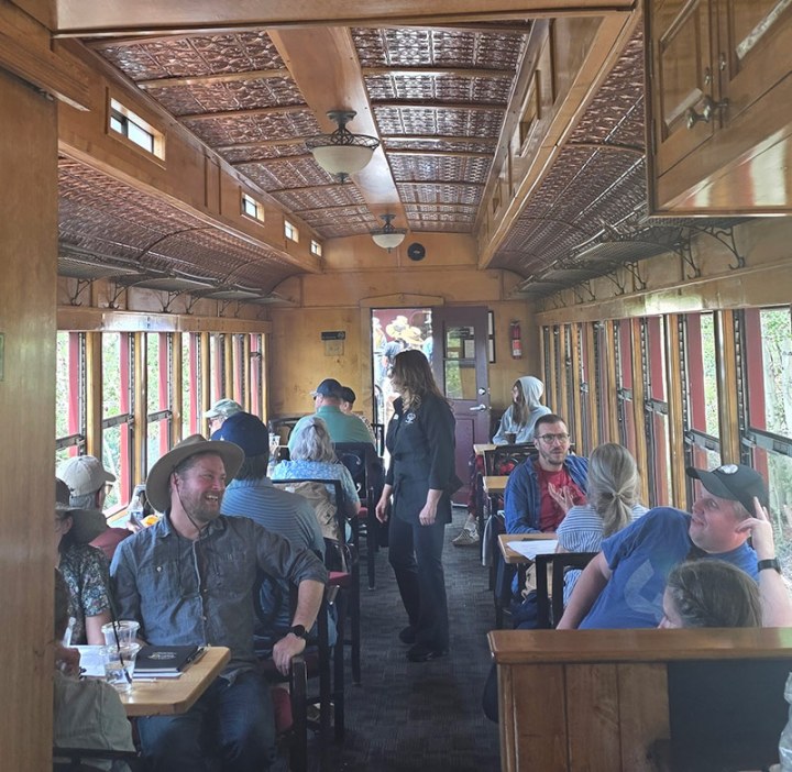 People seated in a vintage train car, with wooden interior and patterned ceiling.