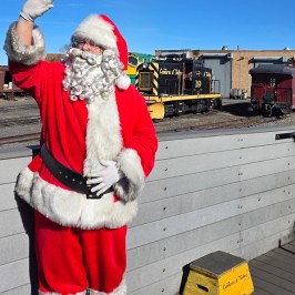 Santa Claus waves on a train platform with a freight train in the background.