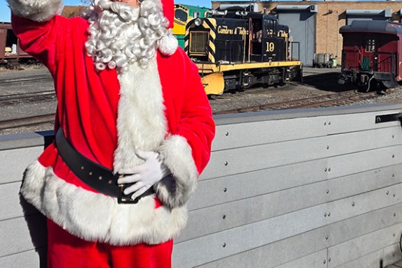 Santa Claus waves on a train platform with a freight train in the background.