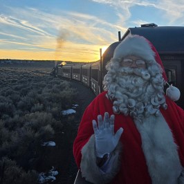 Santa waves from a train with a scenic sunset and open landscape in the background.