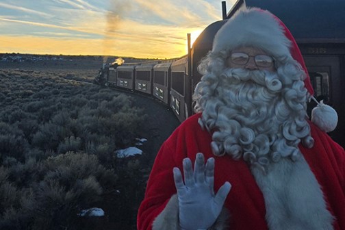 Santa waves from a train with a scenic sunset and open landscape in the background.