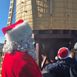 Person in Santa costume looks at wooden water tower on sunny day.