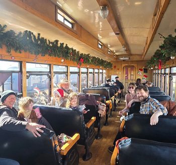 People in festive train car with holiday garlands on windows.